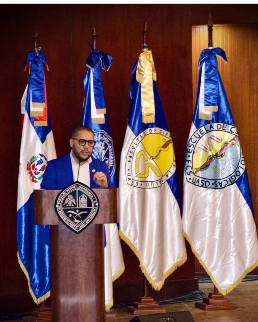 El Dr. Ezequiel Lugo exponiendo en un atril de la Universidad Autónoma de
                    Santo Domingo, ante las banderas de la Facultad de Ciencias de la Salud y de
                    la Escuela de Ciencias Fisiológicas